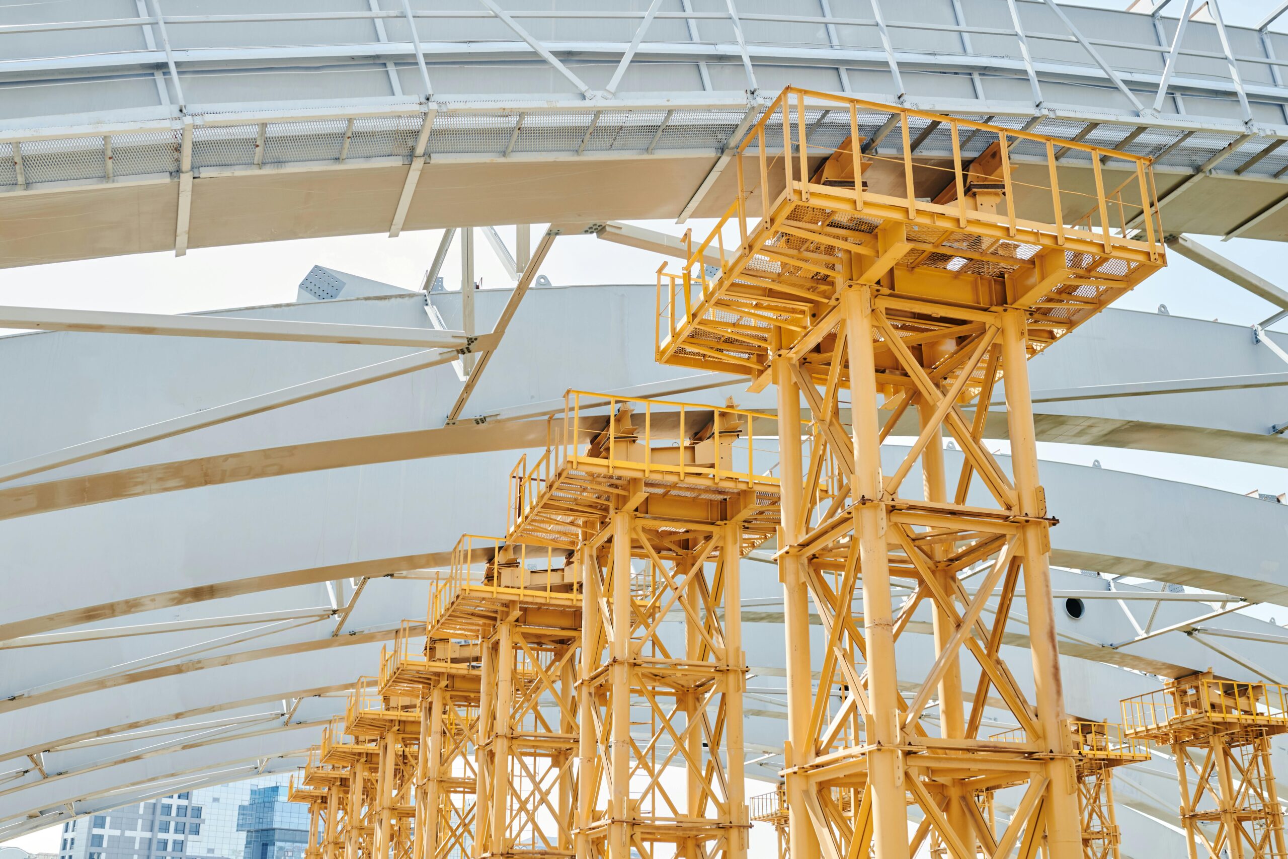 Yellow metal scaffolding beneath steel arches at a construction site under clear daylight.